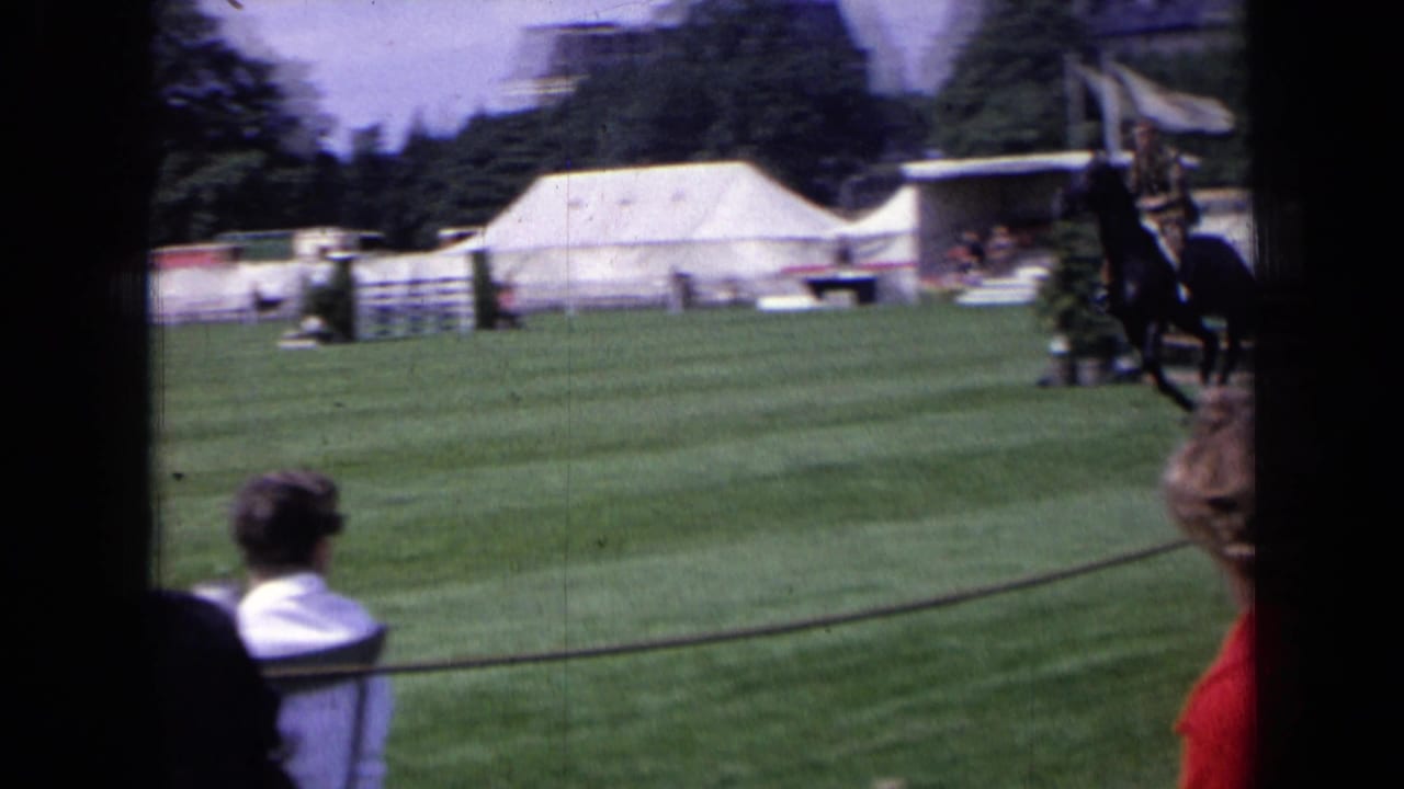 1963 England 8mm frame showing a rider on a dark horse near a show-jumping obstacle with tents, grass, and spectators visible.
