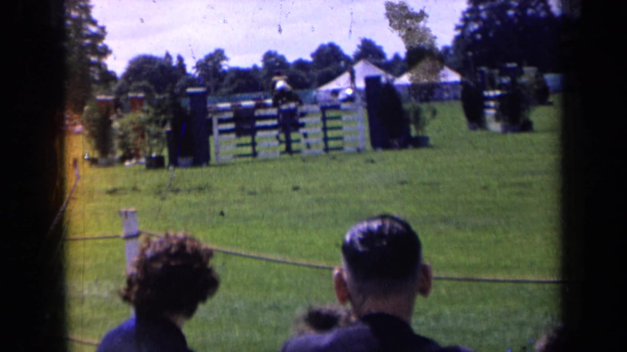 1963 England home-movie frame showing show-jumping obstacles, spectators from behind, tents, and trees around a grass event field.