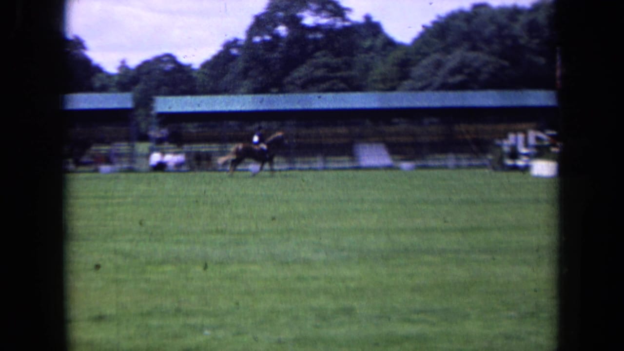 A rider moves across a grassy competition field in England, with spectators and a long covered structure in the background.