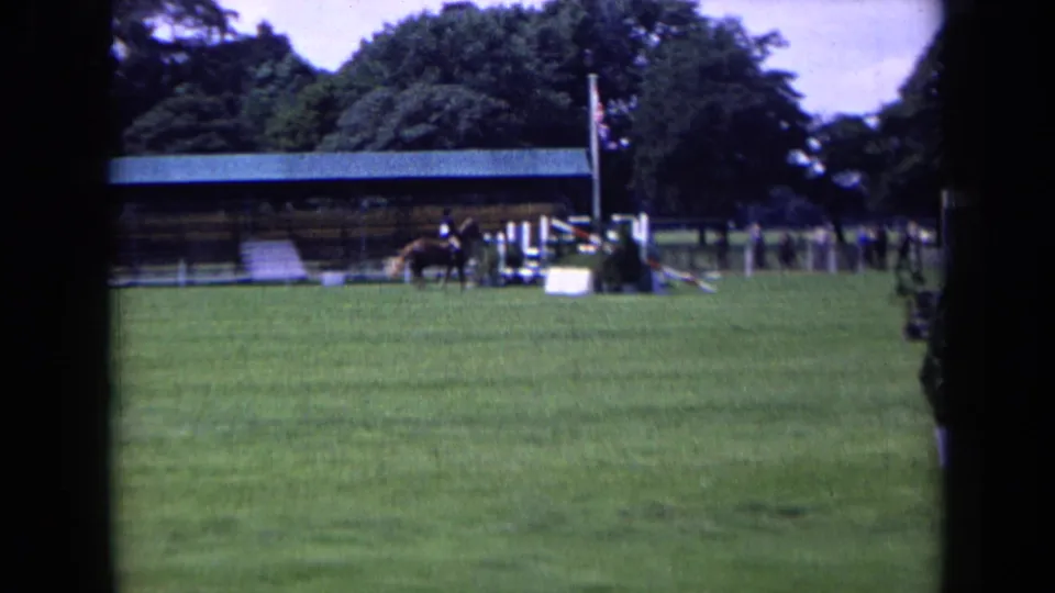 England 1963 frame with a horse and rider near jump obstacles, a covered event structure, trees, and a visible British flag.