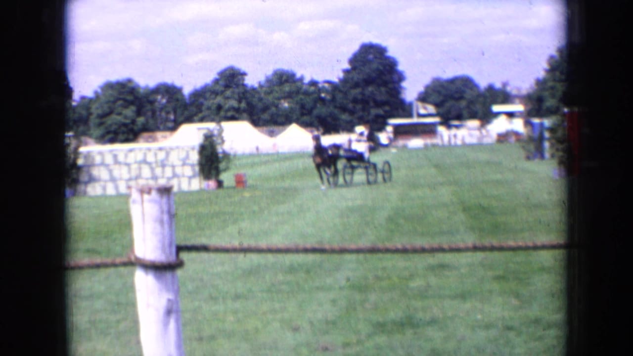 A distant horse-drawn cart or small carriage appears on the English show field near tents, obstacles, trees, and a white foreground post.