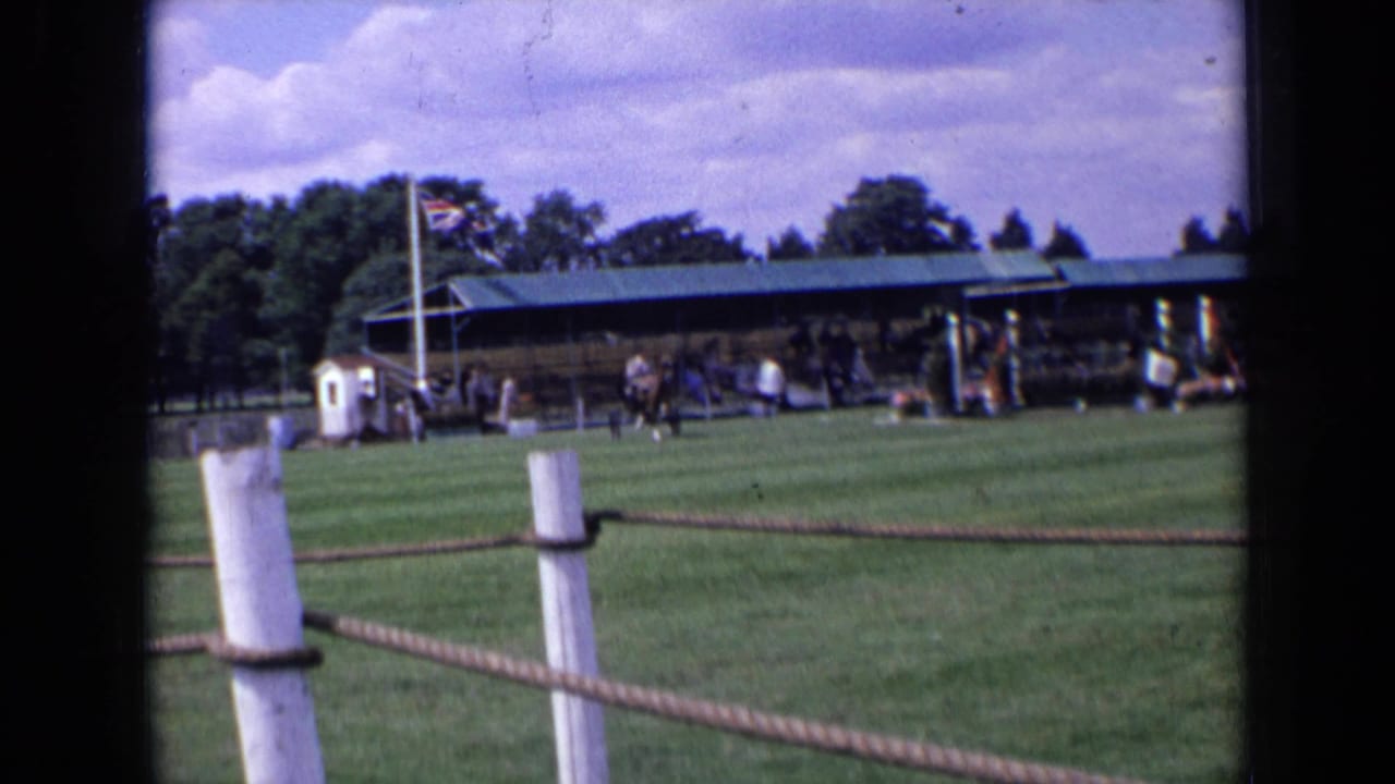1963 England home-movie frame showing a grass field, rope fence, covered spectator stand, Union Jack flag, and horse carriage activity.