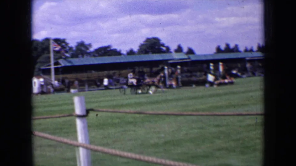 1963 England frame of a horse-drawn carriage crossing a grass field before a covered stand, spectators, rope fencing, and flag.