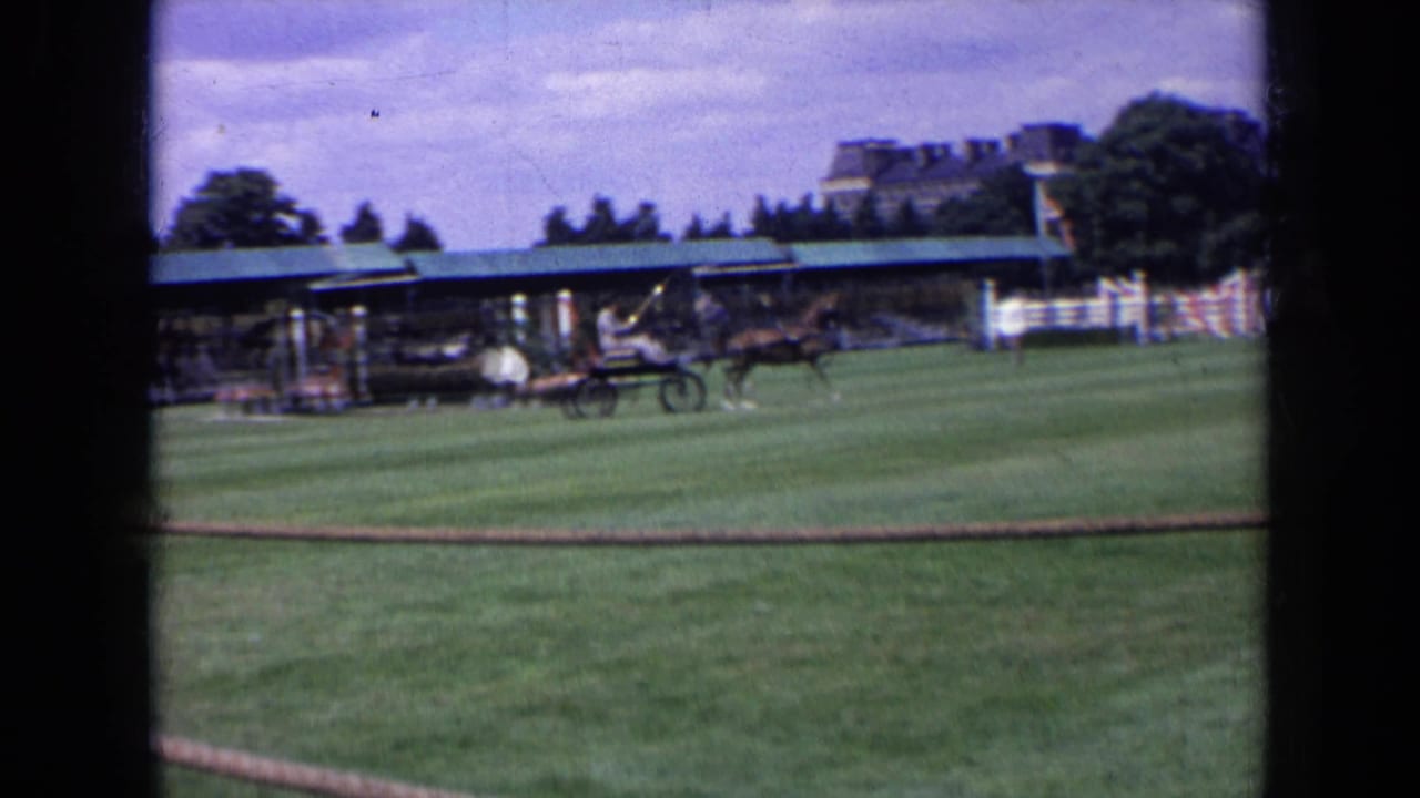 1963 England home-movie frame showing a horse carriage crossing a field near spectators, a covered stand, rope fence, and distant building.
