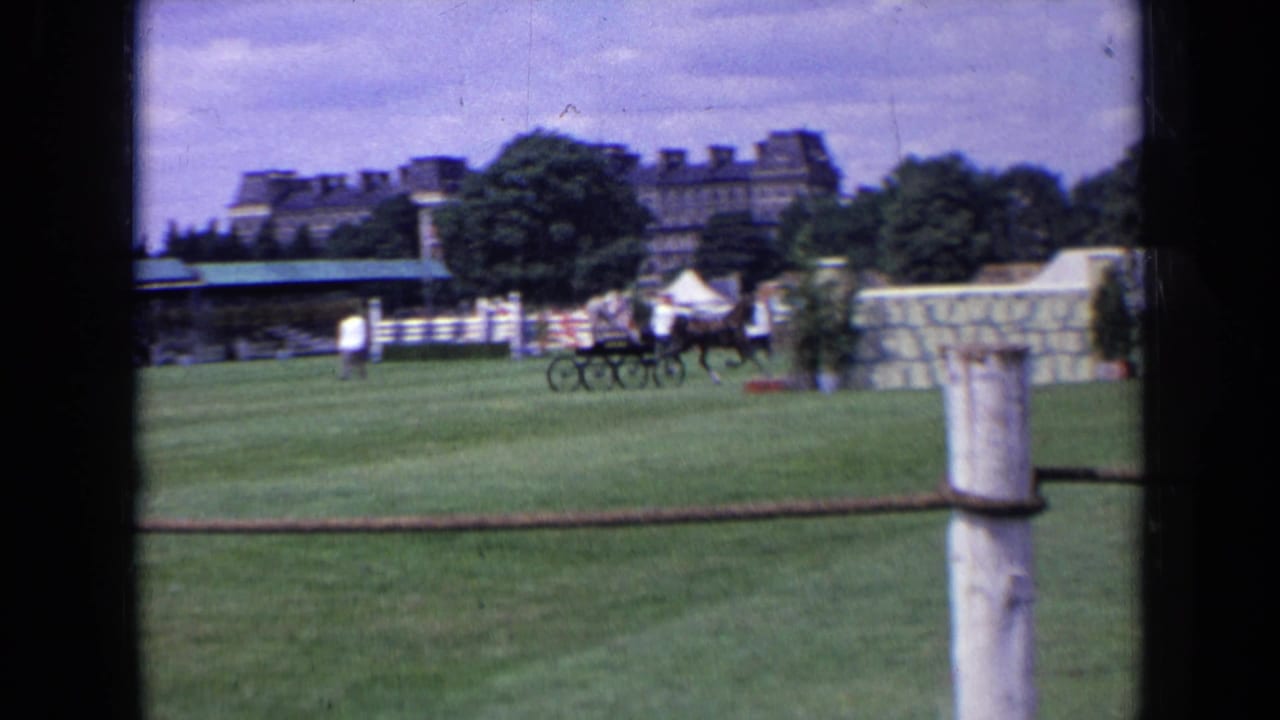 1963 England frame showing a horse-drawn carriage team on a grass field, with rope fencing, trees, tent, spectators, and a large building behind.