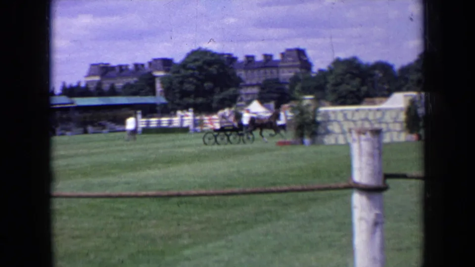 1963 England frame showing a horse-drawn carriage team on a grass field, with rope fencing, trees, tent, spectators, and a large building behind.