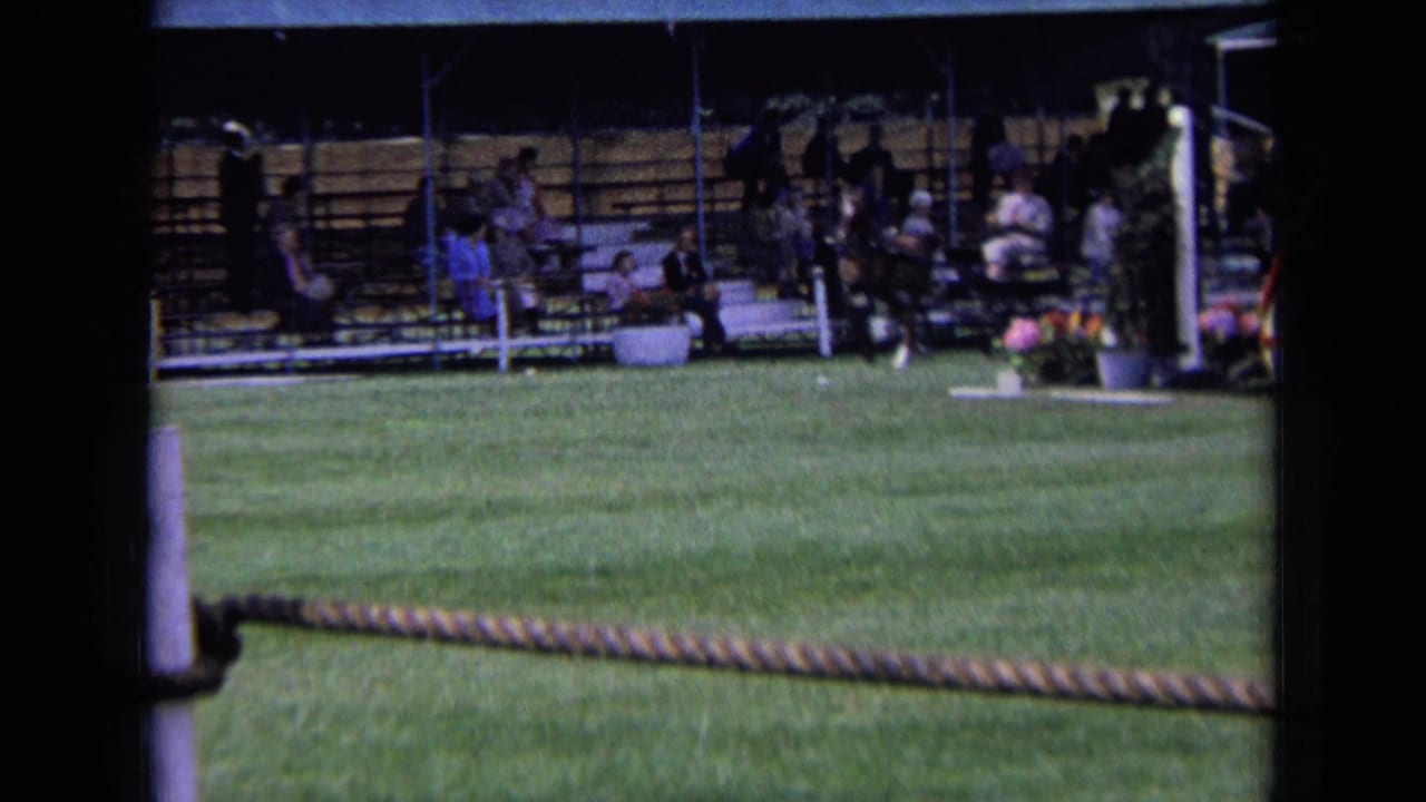1963 England 8mm frame showing a grass ring, rope barrier, posts, and spectators seated under a covered stand.