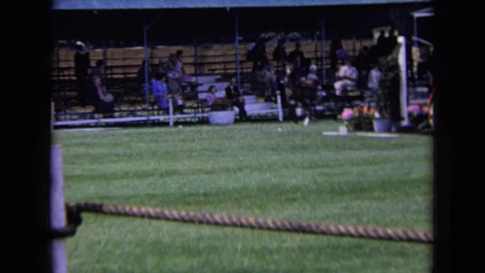 1963 England 8mm frame showing a grass ring, rope barrier, posts, and spectators seated under a covered stand.