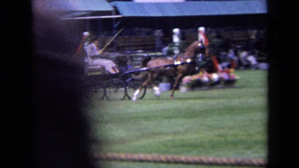 1963 England home-movie frame of a horse-drawn cart crossing a grass field at an equestrian event, with spectators seated in bleachers behind.
