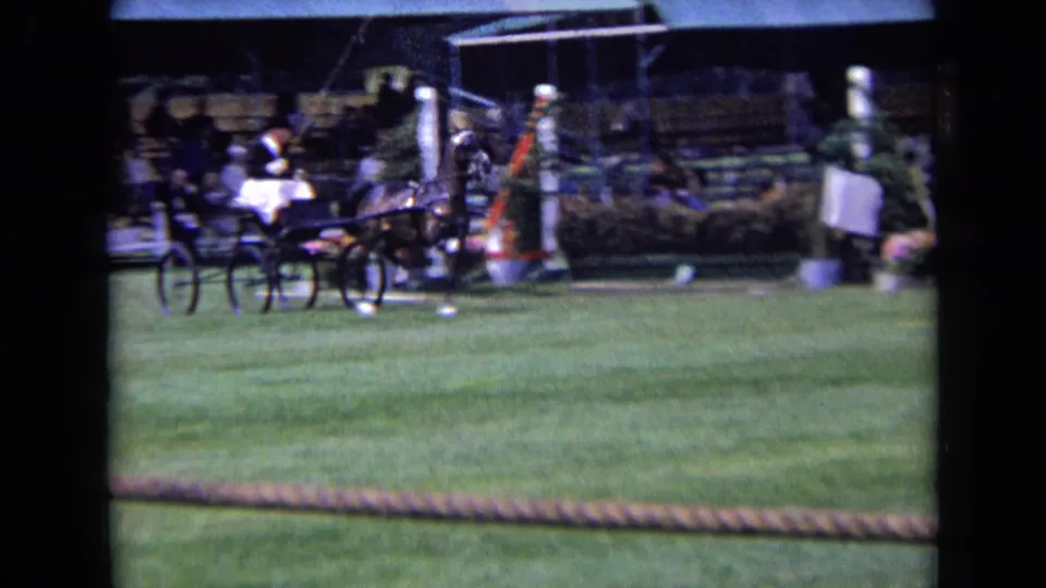 1963 England home-movie frame showing a horse-drawn cart on a grass show field with a driver, barriers and spectators in the background.