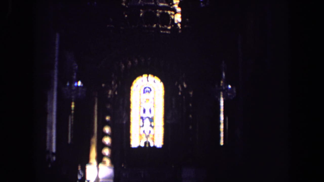 1963 England home-movie frame showing a dim interior with a bright stained-glass window and surrounding dark wall detail.