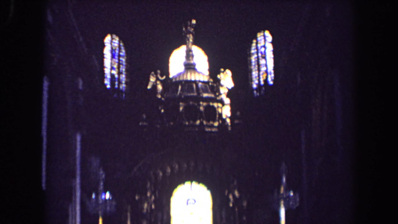 1963 England 8mm frame looking upward at stained glass, dark arches, and gilded angel figures inside a church.