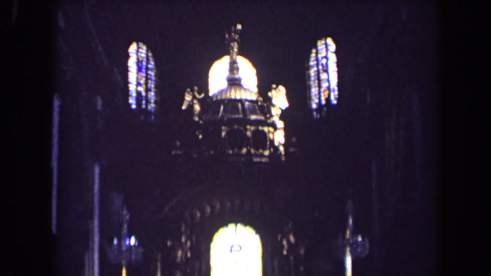 1963 England 8mm frame looking upward at stained glass, dark arches, and gilded angel figures inside a church.