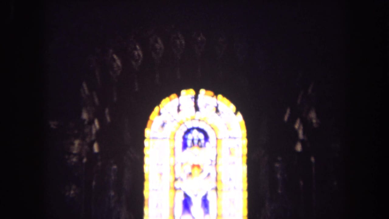 1963 England 8mm frame of a bright stained glass window glowing against the dark interior of a church.