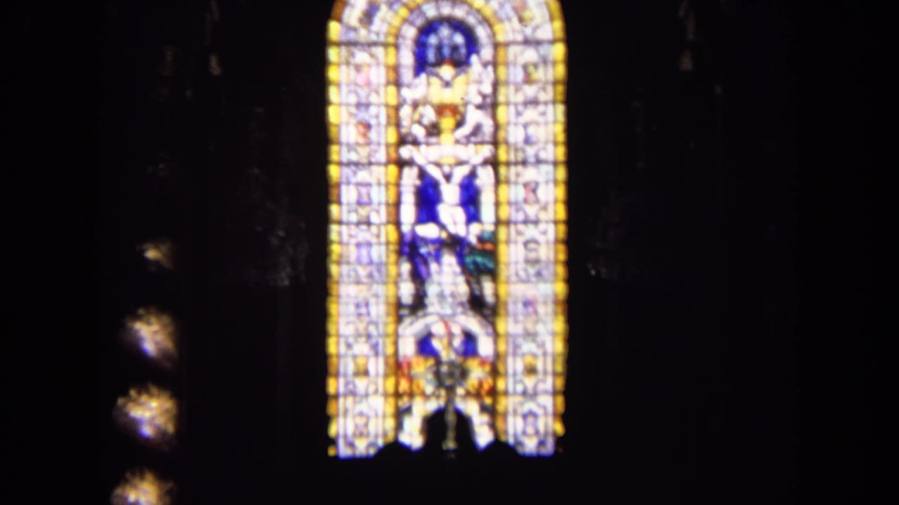 1963 England church interior frame showing a tall stained glass window with a crucifixion-style religious scene in dim surroundings.