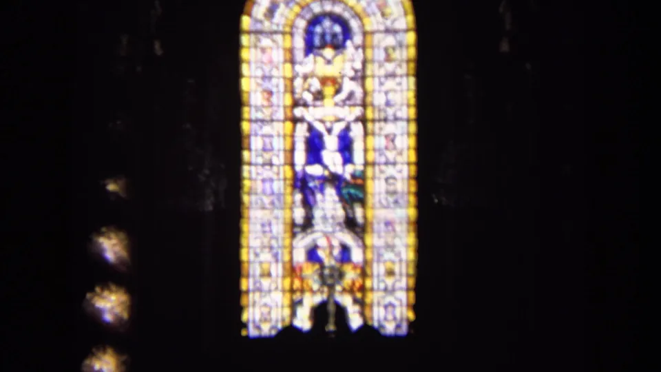 1963 England church interior frame showing a tall stained glass window with a crucifixion-style religious scene in dim surroundings.