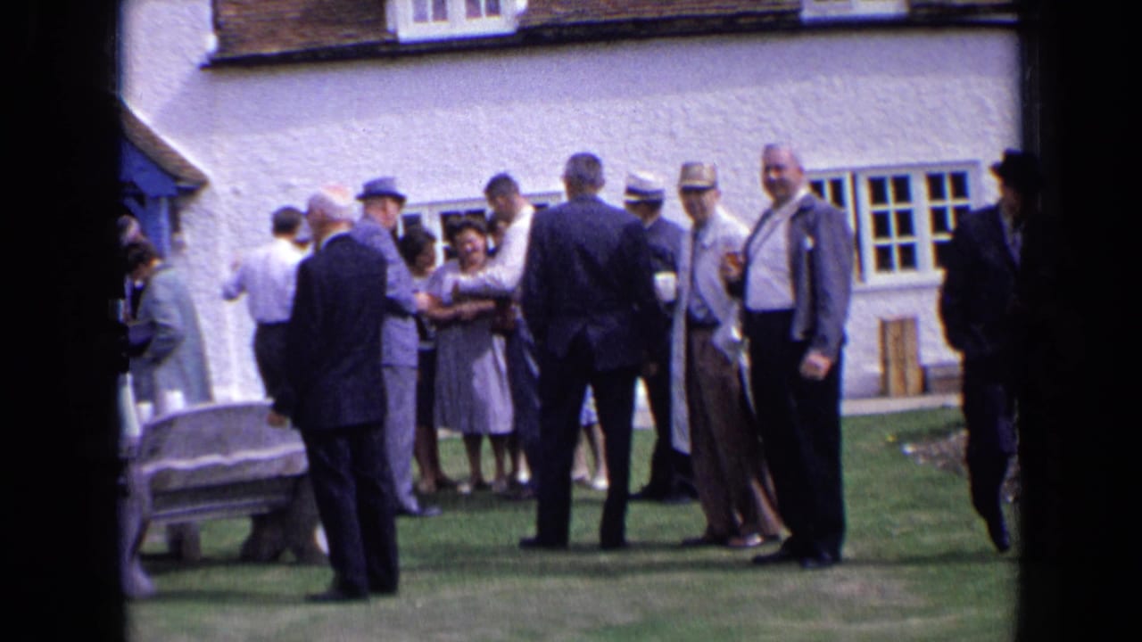 Formally dressed adults gather on a lawn outside a white building with a blue doorway.