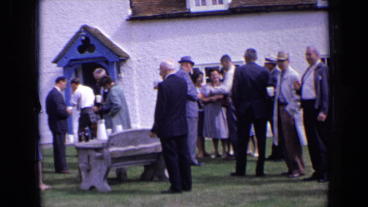 1963 England home-movie frame of formally dressed guests waiting or socializing near a white building, blue door, bottles, and cups.