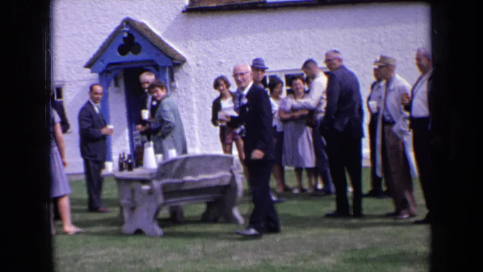 1963 England 8mm still of guests in formal clothing beside bottles, cups, a stone bench, and a blue-trimmed doorway.
