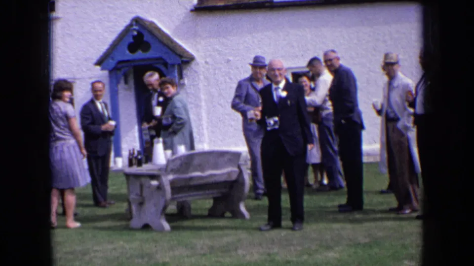 1963 England home-movie frame showing a suited man carrying a camera in front of guests near a blue-doored white building.
