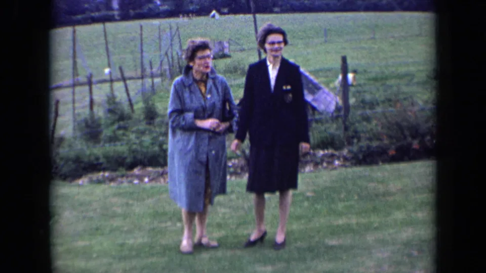 Two adult women in 1960s coats stand together on a lawn in an English countryside garden with fencing behind them.