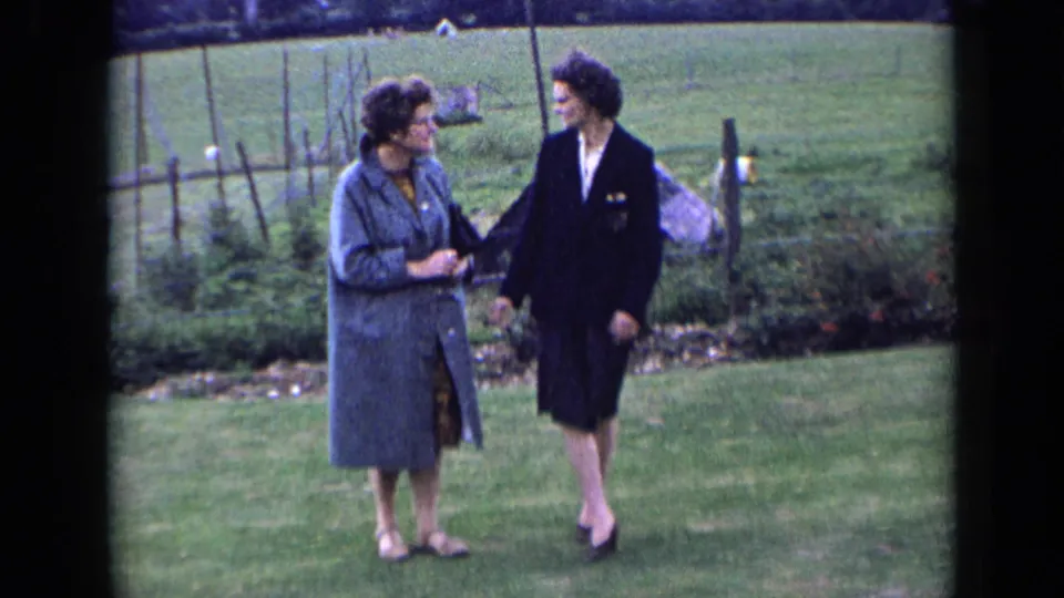 Two women in coats walk together across a lawn in 1963 England, with garden beds, fencing, and open countryside behind them.