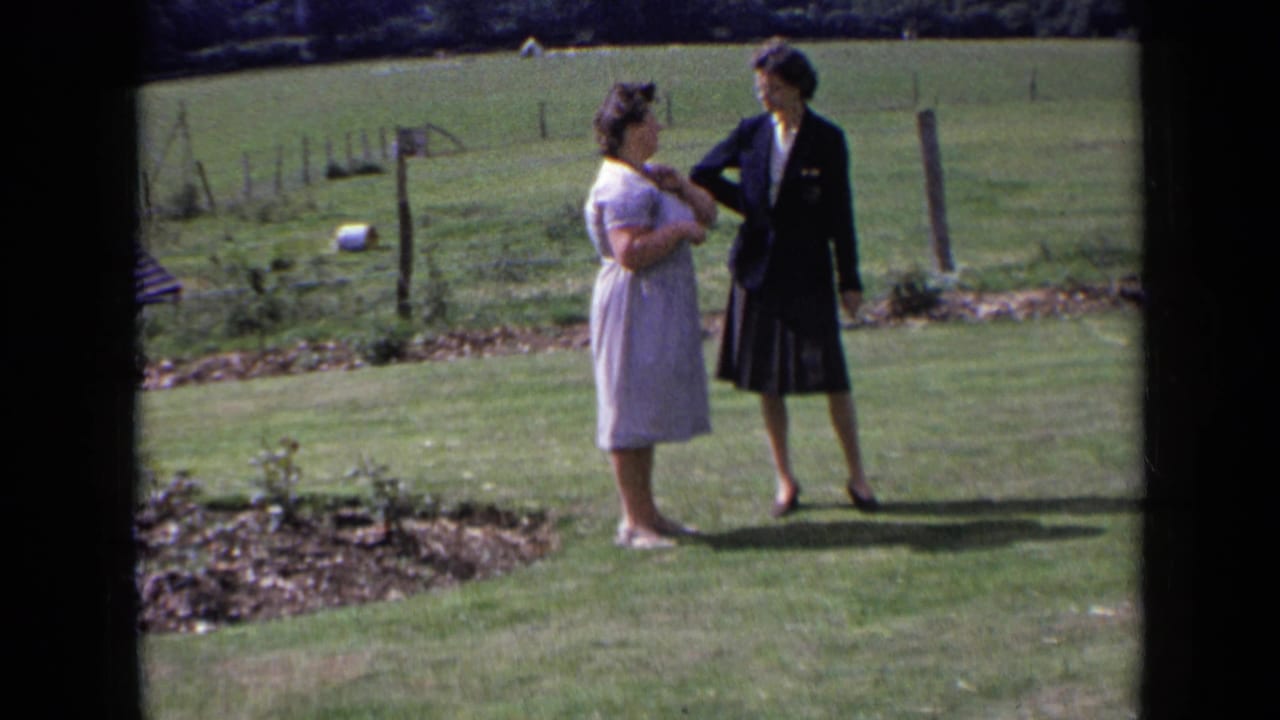 Two adult women in 1963 England stand on a sunlit grassy lawn, one in a pale dress and one in a dark blazer and skirt.
