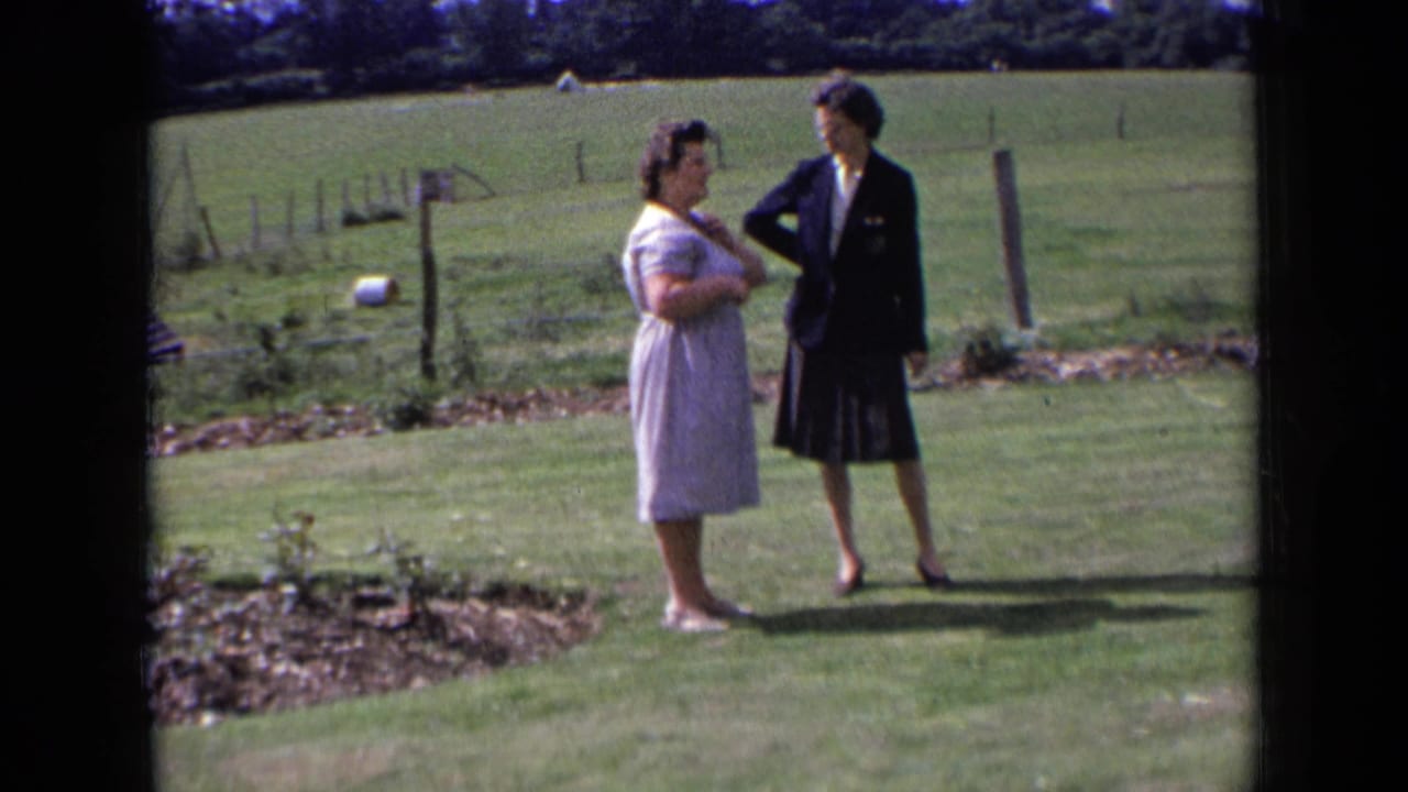Two adult women in rural England stand facing each other on grass, with fence posts and open pasture in the background.