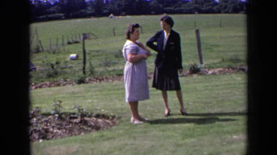 Two adult women in rural England stand facing each other on grass, with fence posts and open pasture in the background.