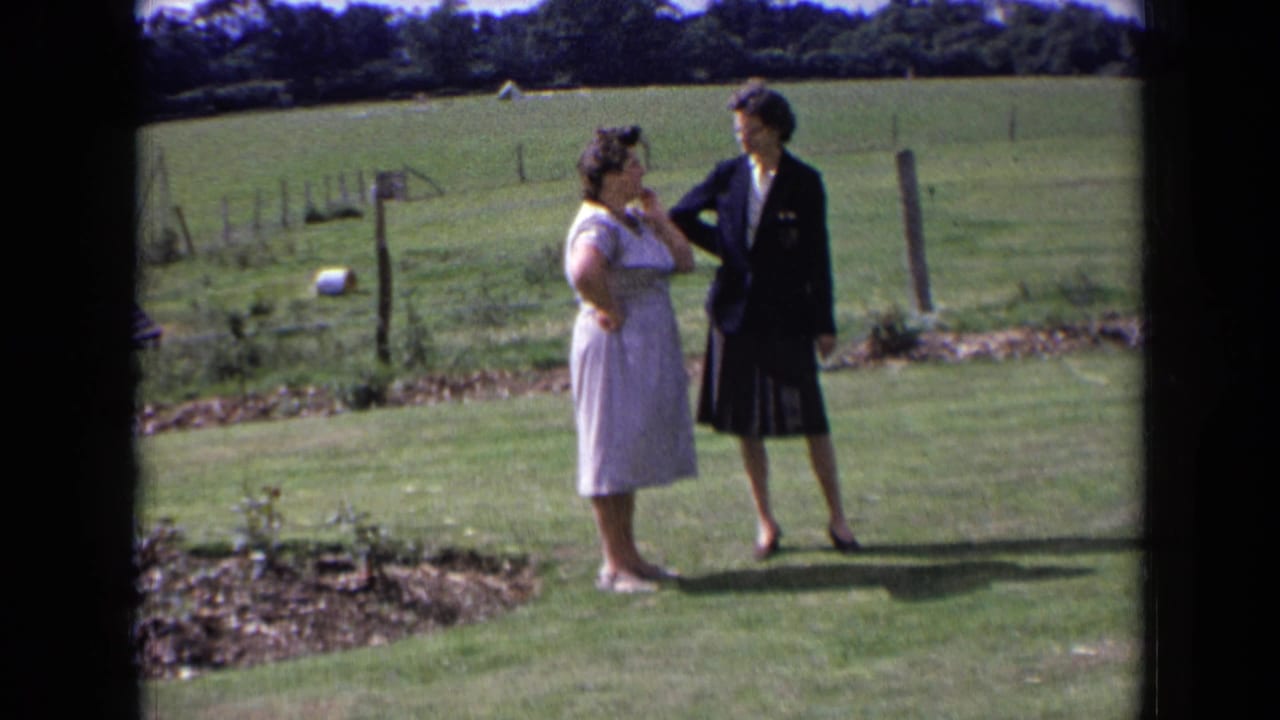 In a 1963 England home-movie frame, two women talk on a lawn while one raises a hand near her face.