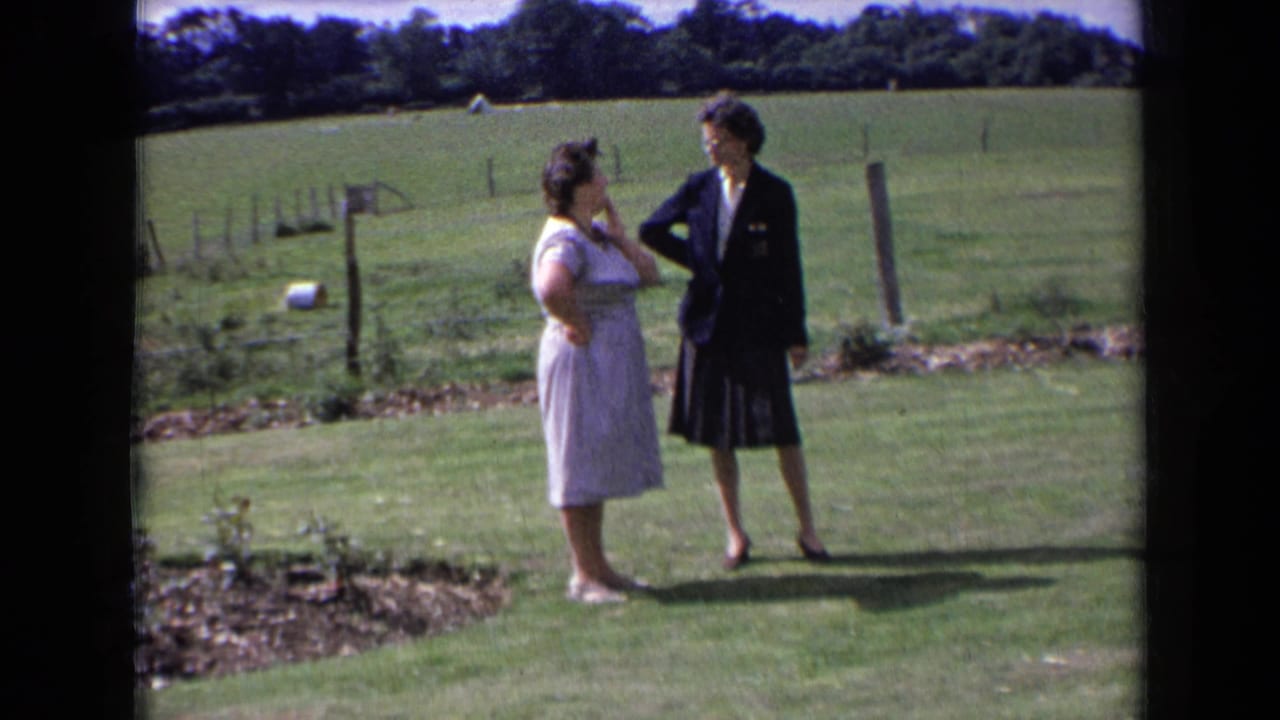 Two adult women in 1963 England face each other on a sunlit lawn, framed by fence posts, pasture, and a dark tree line.