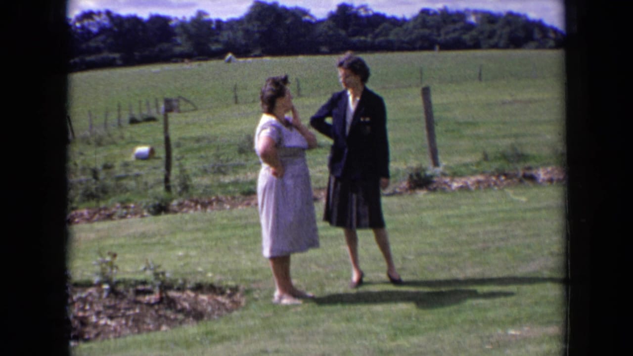 Two women stand on a lawn beside fields and fence posts.