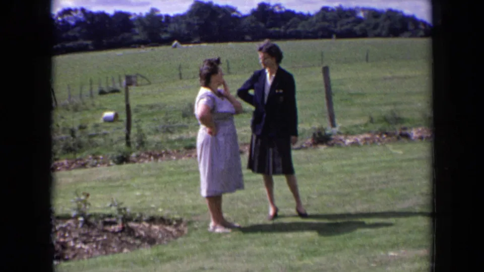 Two women in 1960s England stand in a rural garden, one in a light dress and one in a dark blazer and skirt.
