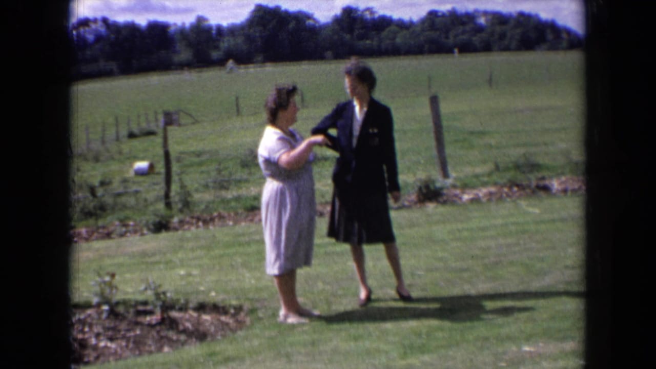 Two women stand in a 1960s England rural garden as one lightly holds or indicates the other's arm while speaking.