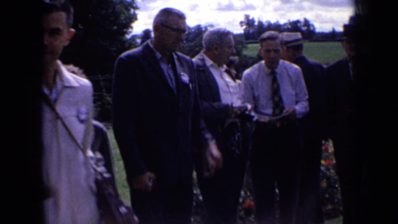 Several adult men in suits and hats gather outdoors in rural England, with garden flowers and fields nearby.