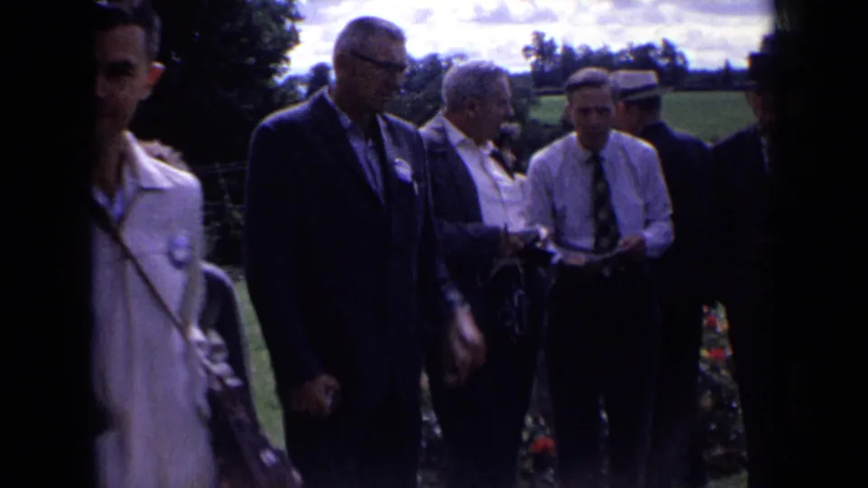 Several adult men in suits and hats gather outdoors in rural England, with garden flowers and fields nearby.