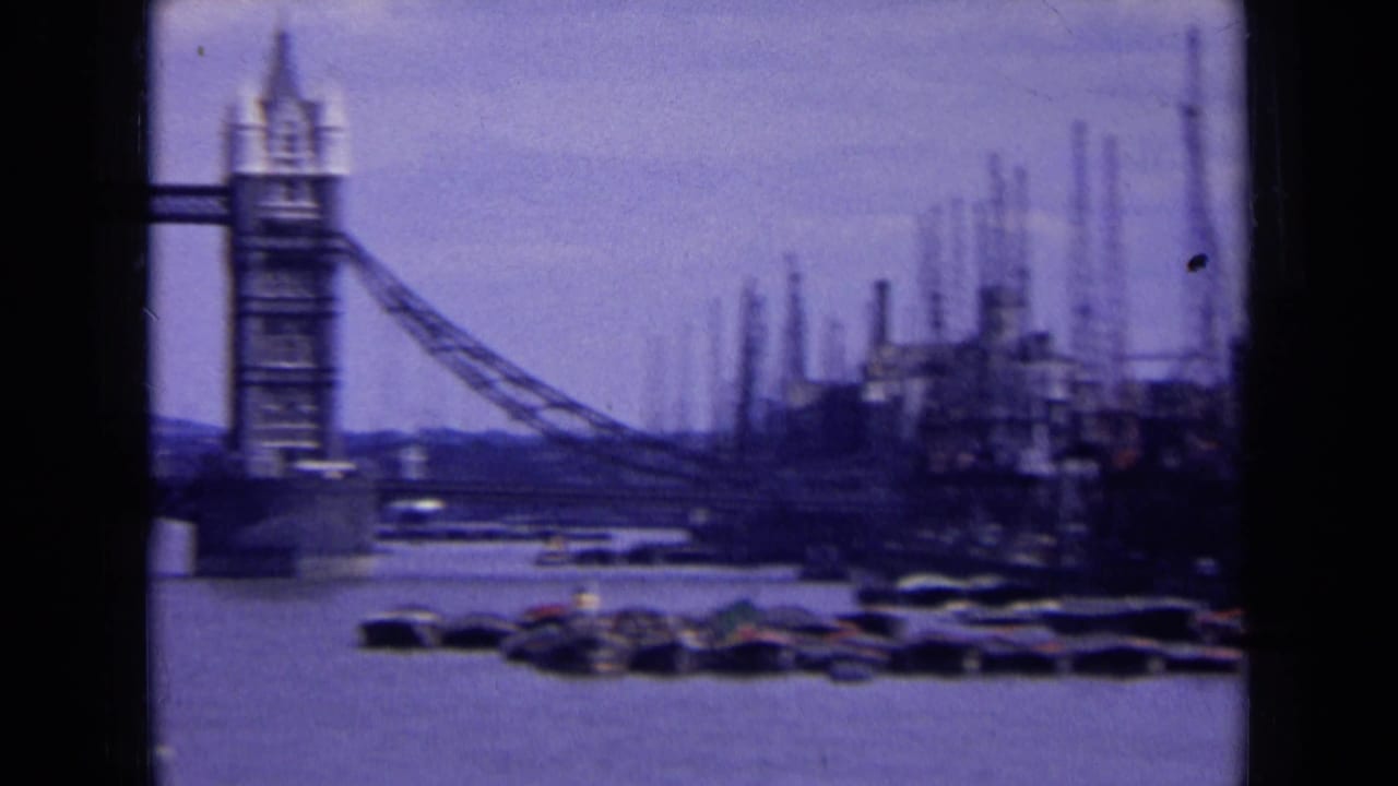 1963 London 8mm frame showing a Tower Bridge tower near riverside cranes, moored boats, and industrial structures on the Thames.