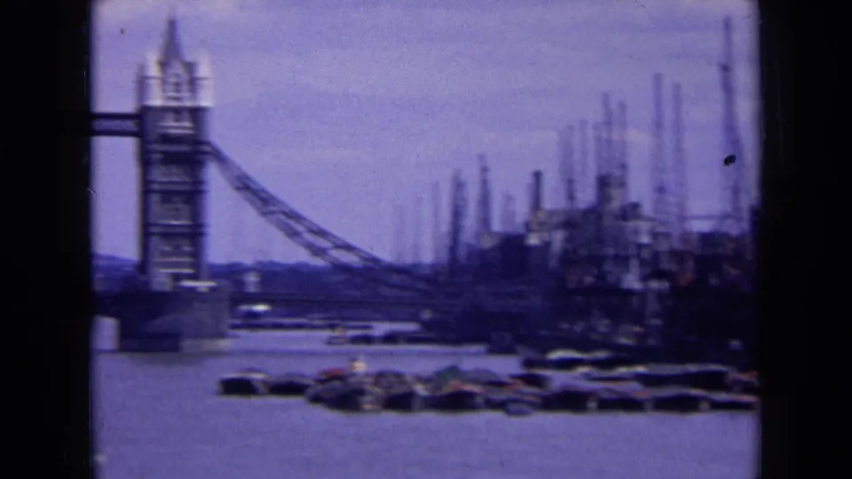 1963 London 8mm frame showing a Tower Bridge tower near riverside cranes, moored boats, and industrial structures on the Thames.