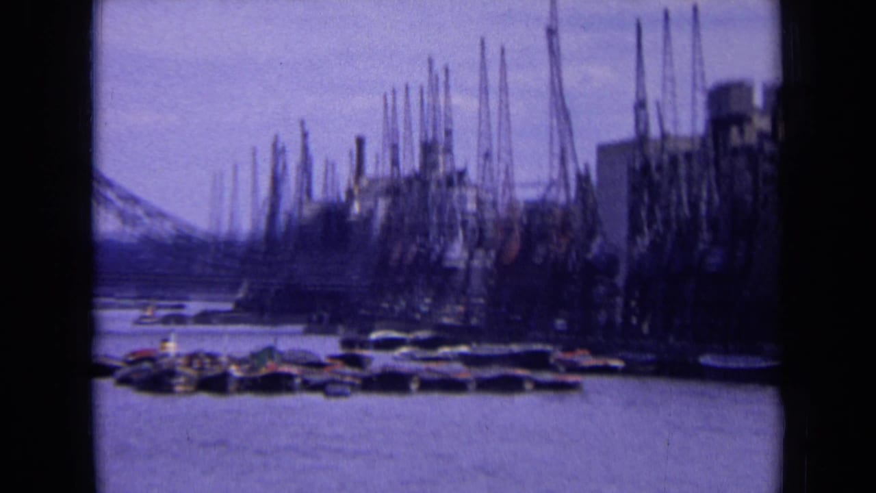 1963 England home-movie frame showing an industrial riverside with cranes, buildings, moored craft, and a distant bridge.