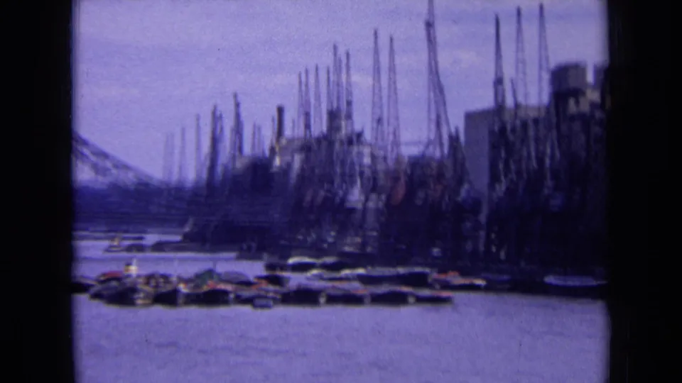 1963 England home-movie frame showing an industrial riverside with cranes, buildings, moored craft, and a distant bridge.