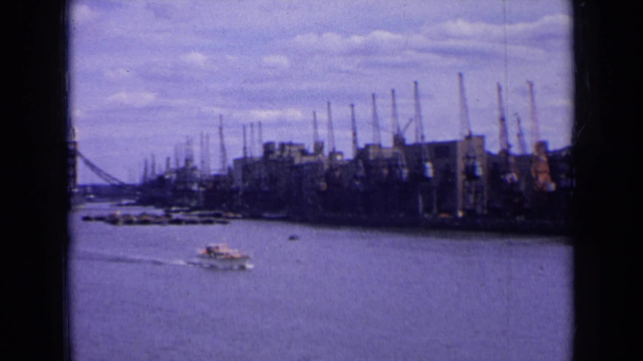 1963 England riverside view with a small boat leaving a wake near industrial cranes, buildings, and a distant bridge.