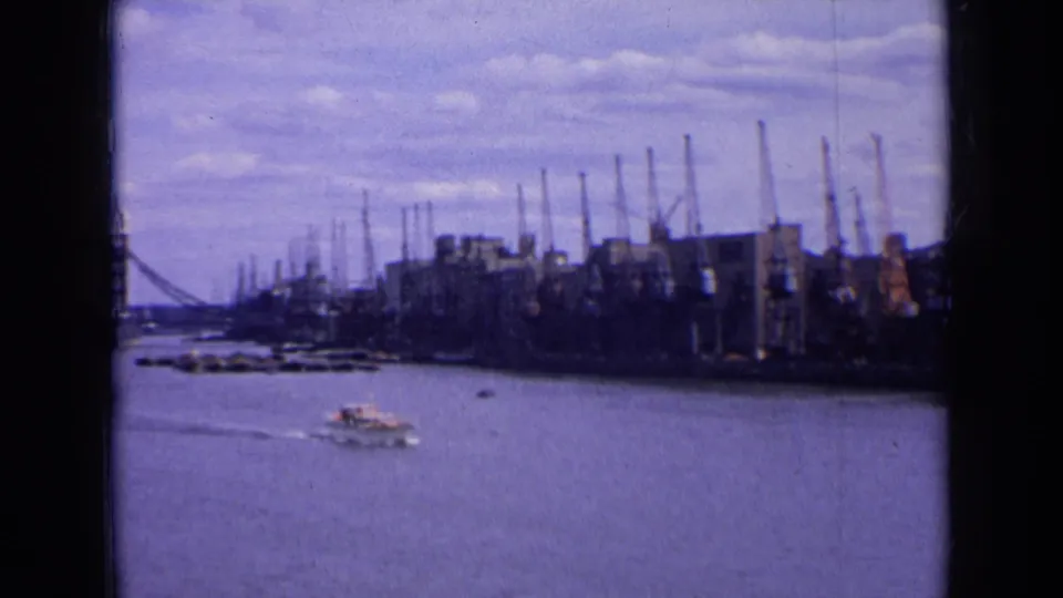 1963 England riverside view with a small boat leaving a wake near industrial cranes, buildings, and a distant bridge.