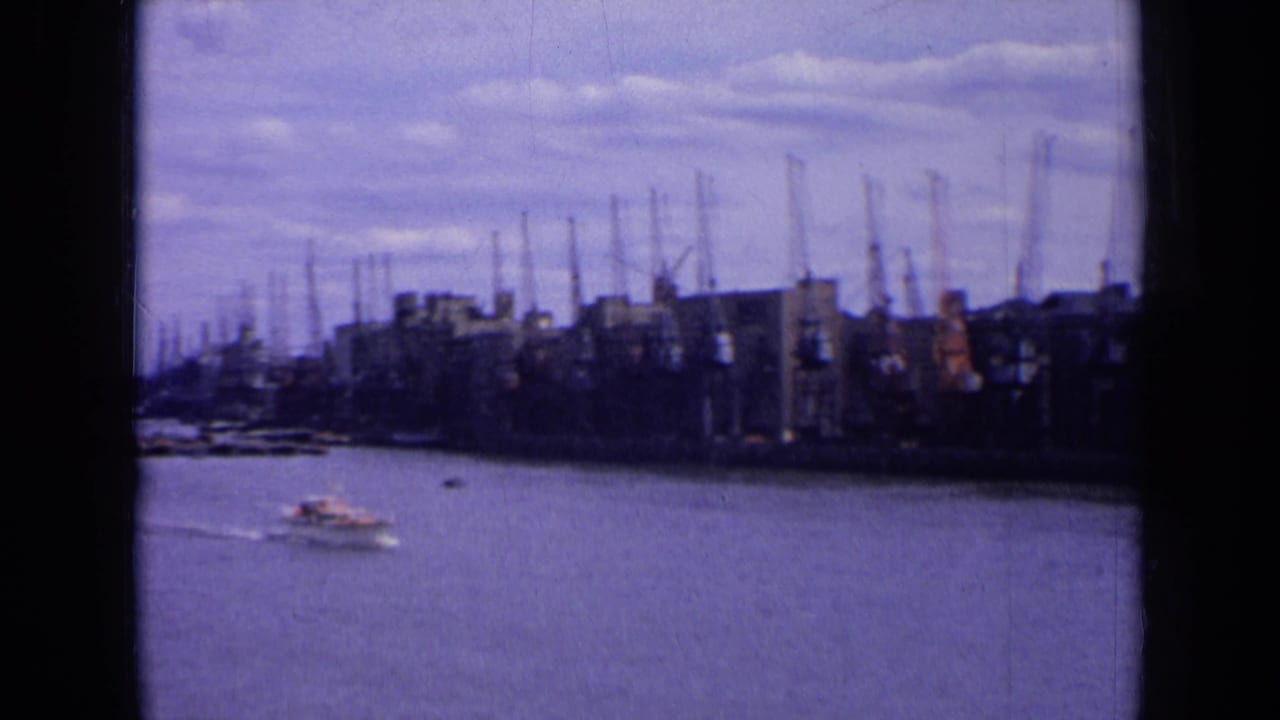 Small boat crossing water in front of distant warehouses and harbor cranes.