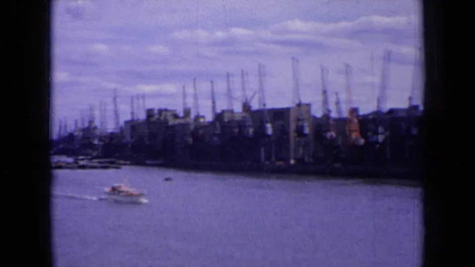 1963 England harbor view with a small boat on the water and distant dockside warehouses backed by many cranes.