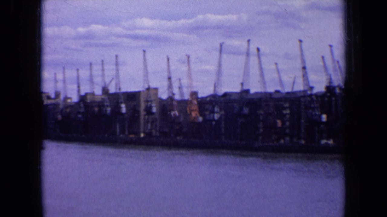 1963 England waterfront seen across the water, with warehouse blocks and numerous harbor cranes under clouds.