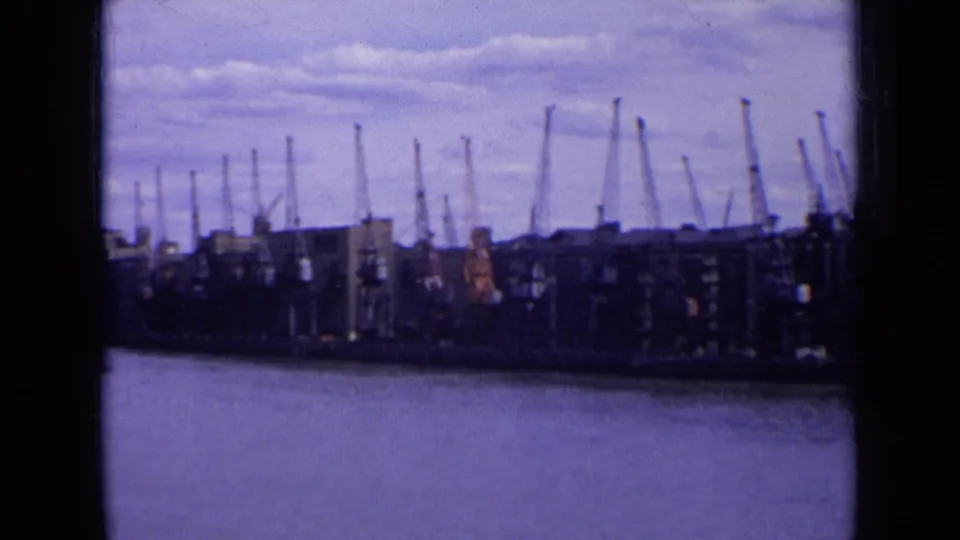 1963 England waterfront seen across the water, with warehouse blocks and numerous harbor cranes under clouds.