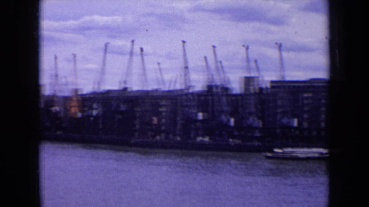 A 1963 England harbor shoreline with cranes rising above dock buildings and a small vessel near the waterfront.