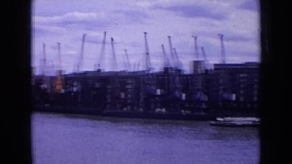 A 1963 England harbor shoreline with cranes rising above dock buildings and a small vessel near the waterfront.