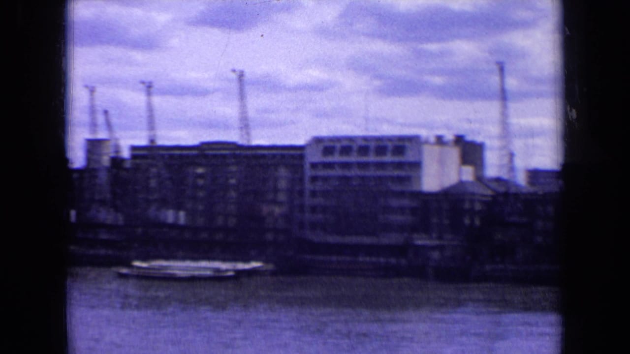 Riverside buildings and cranes seen across water under a cloudy sky.