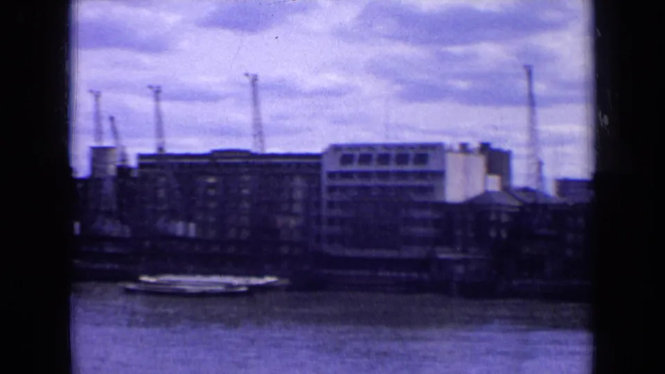 1963 England 8mm frame of an industrial riverfront with cranes, warehouses, water, and cloudy sky, framed by dark film edges.